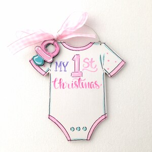 May include: A white onesie-shaped Christmas ornament with the text "MY 1st Christmas" in pink and blue. It has a pink pacifier charm and a pink gingham ribbon for hanging. The ornament is decorated with pink polka dots.