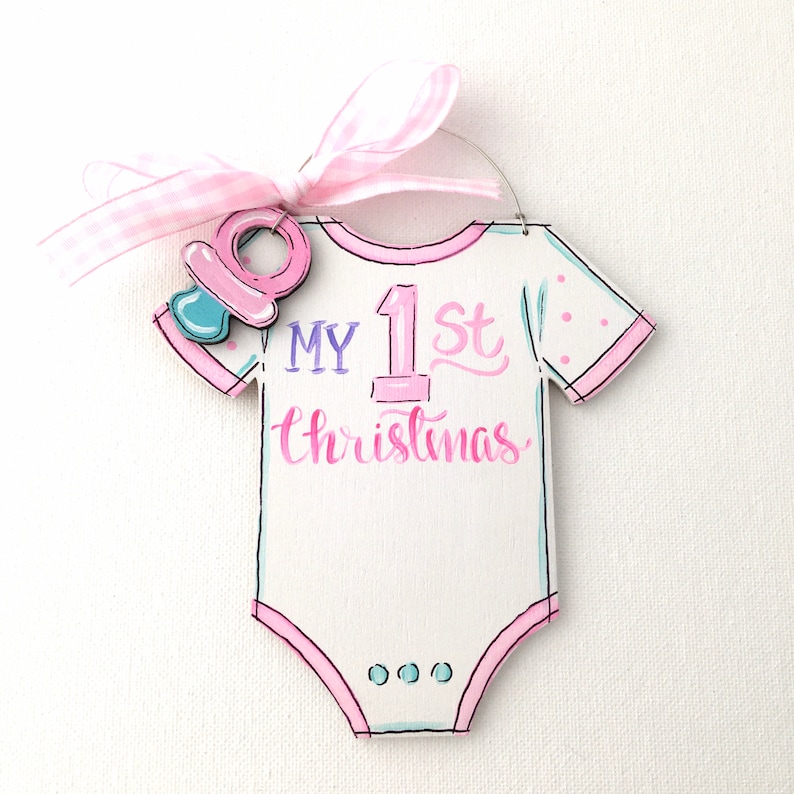 May include: A white onesie-shaped Christmas ornament with the text "MY 1st Christmas" in pink and blue. It has a pink pacifier charm and a pink gingham ribbon for hanging. The ornament is decorated with pink polka dots.