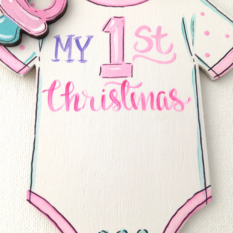May include: A wooden baby onesie ornament with the text "MY 1st Christmas" in pink and purple script. The onesie is white with pink and teal accents. The ornament has a pink and teal design at the top left.