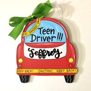 May include: A red, car-shaped wooden sign with the text "Teen Driver!!!" and the name "Jeffrey" in black script. The sign has yellow caution tape with "KEEP BACK!" and "DANGER!" written on it, and is adorned with a green ribbon.