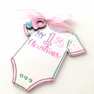 May include: A white wooden baby onesie ornament with pink trim and the text "MY 1st Christmas" in pink and blue. A pink pacifier and a pink and white gingham ribbon are attached to the ornament.