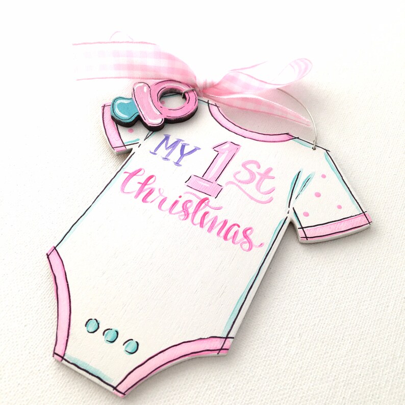 May include: A white wooden baby onesie ornament with pink trim and the text "MY 1st Christmas" in pink and blue. A pink pacifier and a pink and white gingham ribbon are attached to the ornament.