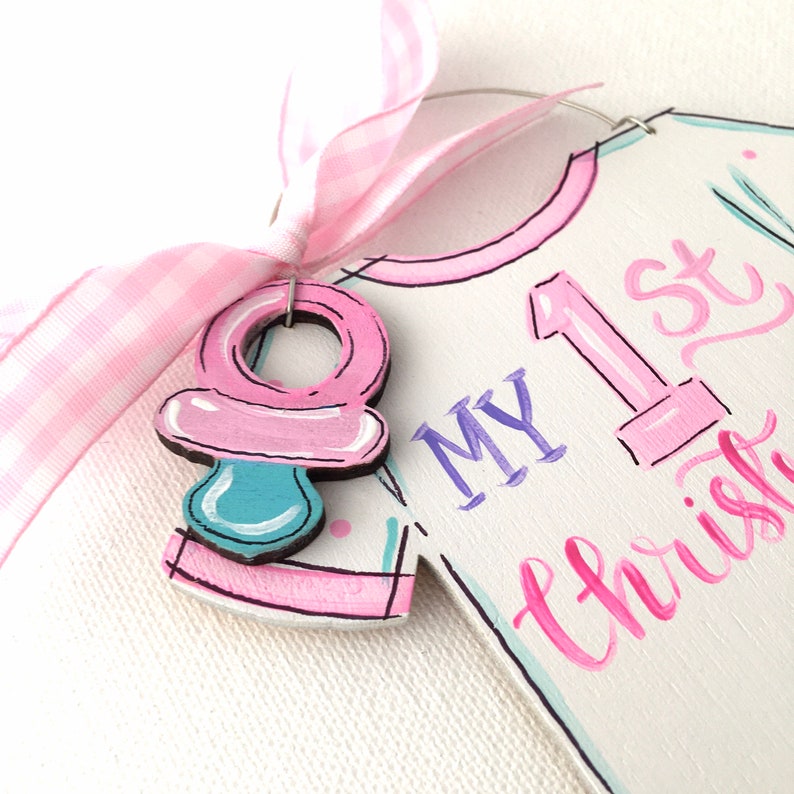 May include: A wooden baby ornament with a pink pacifier charm and a pink gingham ribbon. The ornament is shaped like a onesie and has the words "My 1st Christmas" in pink and purple.