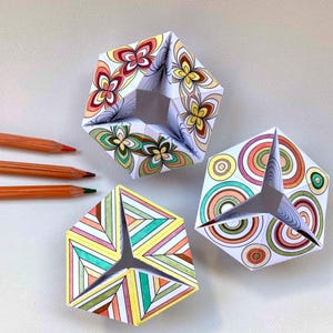 May include: Three folded paper geometric shapes with intricate hand-colored patterns in shades of orange, yellow, green, and brown, accompanied by three orange-colored pencils. The shapes feature floral, circular, and striped designs.