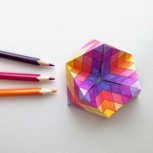 Może przedstawiać: Kolorowa geometryczna rzeźba z papieru z otworem w kształcie gwiazdy. Trzy kredki w kolorze fioletowym, różowym i pomarańczowym umieszczone są w pobliżu rzeźby. Kredki są zaostrzone. Rzeźba jest wykonana z papieru i wielokolorowa.