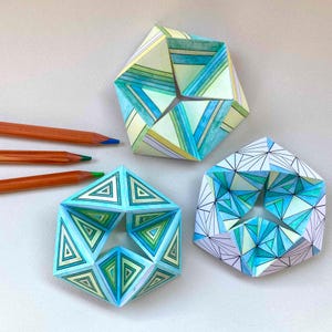May include: Three geometric paper sculptures with intricate hand-drawn patterns in shades of blue, green, and yellow, accompanied by three orange-coloured pencils. The sculptures feature complex folded triangular designs and star-shaped centres.