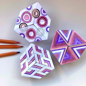 May include: Three folded paper geometric shapes with intricate hand-drawn patterns in shades of purple, pink, and brown. One shape features concentric circles, another displays triangular motifs, and the third shows a chevron pattern. Three colored pencils are placed to the left.