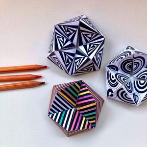 May include: Three geometric paper sculptures with intricate patterns. One features black and white triangles, another black and white concentric circles, and the third multicolored stripes. Three wooden pencils are positioned to the left.