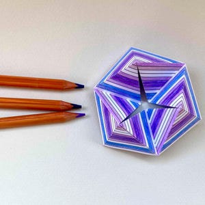 May include: Three wooden pencils with orange casings and dark blue leads are positioned horizontally next to a geometric paper sculpture. The sculpture features a repeating pattern of purple, blue, and white stripes arranged in a star-like shape.