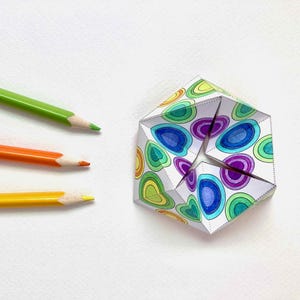 May include: A folded paper geometric shape with a colorful pattern of layered ovals in shades of blue, green, yellow, and purple. Three colored pencils in green, orange, and yellow are arranged on a white surface.