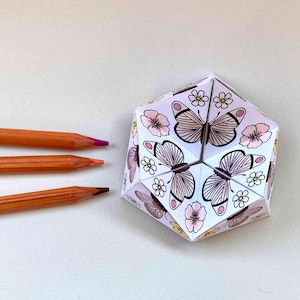 May include: A hexagonal paper craft object featuring black-lined illustrations of butterflies and flowers, partially colored in shades of pink and yellow. Three wooden-cased colored pencils with orange barrels are positioned to the left of the object.