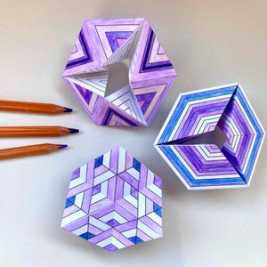 Puede incluir: Tres esculturas geométricas de papel con intrincados patrones dibujados a mano en tonos morados, azules y blancos. Dos esculturas son hexagonales con centros abiertos, mientras que la tercera es una forma hexagonal plana y con mosaicos.