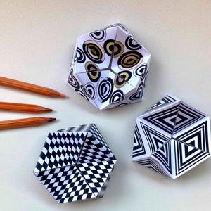 May include: Three hexagonal paper objects with geometric patterns. One features black, white, and gold concentric circles. Another displays a black and white checkered grid. The third shows black and white nested triangles. Three orange pencils are included.