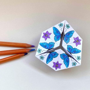 May include: A folded paper fortune teller featuring hand-colored illustrations of blue butterflies and purple and teal flowers. Three wooden colored pencils with orange casings and blue leads are positioned to the left of the paper craft.