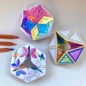 May include: Three geometric paper structures with intricate hand-drawn patterns. One is a solid hexagonal shape with a rainbow star design. Another is a hollow hexagonal ring with floral motifs. The third is a hollow geometric ring with striped patterns.