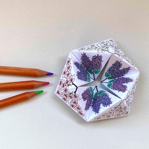 May include: A paper fortune teller featuring intricate black and white line art of floral clusters, with some sections partially colored in shades of purple and pink. Three wooden colored pencils with purple, pink, and green tips lie nearby.