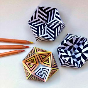 May include: Three geometric paper sculptures with intricate patterns. One features black and white stripes, another has a black and white checkered design, and the third displays concentric triangles in shades of yellow, orange, and black, alongside three orange pencils.