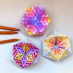 May include: Three hexagonal paper folding toys with intricate geometric patterns in shades of purple, pink, orange, and yellow. Two of the toys feature angular chevron designs, while the third displays overlapping circular line art. Three pencils.