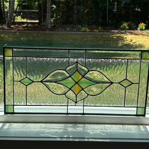 May include: Rectangular stained glass window with a black frame. The design features green, yellow, and clear glass panels. The central design includes a stylized floral motif. The window is resting on a white windowsill.