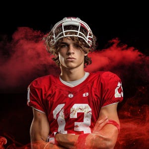 May include: A young man wearing a red football jersey with the number 13 on it. He is wearing a white helmet with a face mask and has his arms crossed. He is standing in front of a red smoke background.