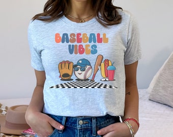 May include: A light blue t-shirt with the text "BASEBALL VIBES" in rainbow colours. The shirt features a cartoon baseball, glove, bat, hot dog, and drink.