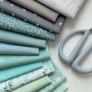 May include: A stack of folded fabric pieces in various shades of gray, teal, and mint green, featuring patterns such as white crosses, delicate floral outlines, small polka dots, geometric chevrons, and abstract watercolor splashes, next to gray scissors.
