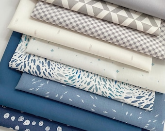 May include: A stack of folded fabric pieces in various shades of blue, gray, and white. Patterns include geometric triangles, a small gray gingham check, scattered stars, a school of blue fish, small white birds, and white circular scribbles.