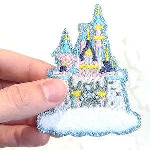 May include: A blue and pink embroidered patch of a fairytale castle with white clouds. The castle has yellow accents and a gray gate.