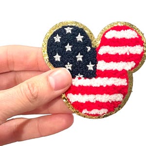 May include: A red, white, and blue American flag in the shape of a Mickey Mouse head with a gold glitter outline.