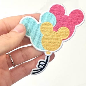 May include: Embroidered patch featuring three Mickey Mouse-shaped balloons in pastel colors: light blue, pink, and yellow. The balloons are outlined in white with black and white details for the string and bow. A fun, decorative item.