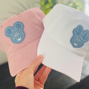 May include: Two baseball caps with embroidered cartoon characters. One cap is pink with a blue Mickey Mouse design. The other cap is white with a blue Minnie Mouse design.