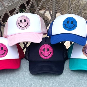 May include: Six trucker hats with white mesh backs and different colored fronts. Each hat features a large, glittery smiley face patch in a different color. The colors are pink, red, blue, navy, teal, and pink.