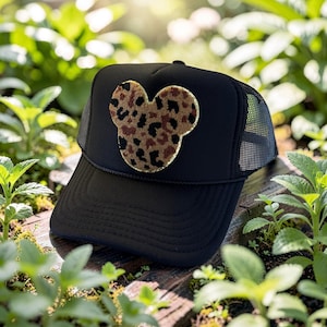 May include: Black trucker hat with a mesh back and a curved brim. The front of the hat features a Mickey Mouse head shape with a leopard print design in brown, black, and tan. The hat is sitting on a wooden surface surrounded by green plants.