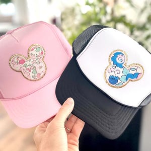 May include: Two trucker hats, one pink and one black, each featuring a Mickey Mouse-shaped floral design. The pink hat has a matching pink mesh back, while the black hat has a white front and black mesh back.