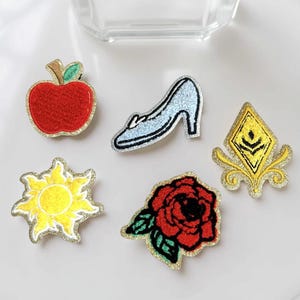 May include: A collection of embroidered patches featuring fairytale-themed designs. Includes a red apple, a blue high-heeled shoe, a yellow sun, a red rose, and a gold and yellow emblem. Each patch has a glittery gold border.