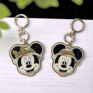 May include: Two gold-colored keychains featuring Mickey and Minnie Mouse in safari hats. The character faces are black, white, and tan, with a gold glitter border. Each keychain has a gold ring for attachment.