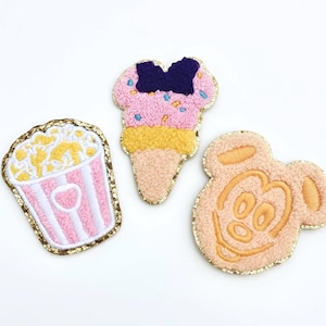 May include: Three embroidered patches: a popcorn bucket with pink and white stripes, a pink ice cream cone with a blue bow, and a Mickey Mouse head. All patches have gold glitter borders.