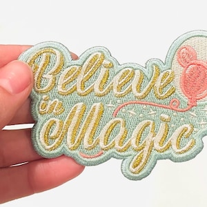 May include: A light blue embroidered patch with gold lettering that says "Believe in Magic". A pink balloon with mouse ears is attached to the lettering.