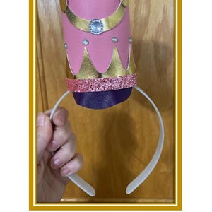Puede incluir: Sombrero de diadema de cascanueces rosa y dorado con una diadema blanca. El sombrero presenta adornos dorados, una banda rosa brillante y un acento de joya. El texto "Nutcracker Headband Hat" está en la parte inferior de la imagen.
