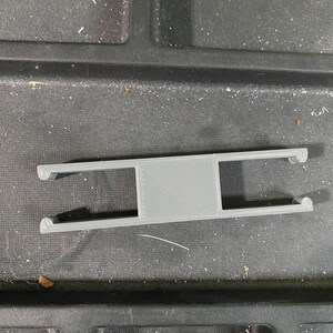 May include: A gray 3D-printed object, rectangular in shape with two parallel bars and a central square. The ends of the bars curve upwards. The object is likely a component or part of a larger assembly.