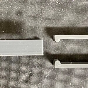 May include: Two gray 3D-printed plastic components. One is a rectangular bar, and the other is a rectangular frame with curved ends. The frame has a textured edge. Both pieces are on a dark, textured surface.