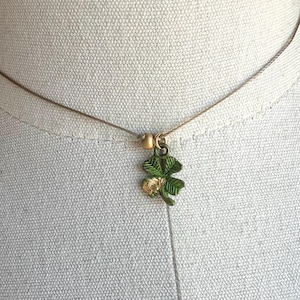 May include: A delicate necklace featuring a small, green and gold shamrock pendant. The four-leaf clover charm hangs from a thin, neutral-colored cord. The pendant has green enamel leaves with gold accents, creating a subtle, elegant accessory.