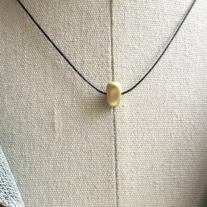 May include: A minimalist necklace featuring a small, oblong, cream-colored bead suspended on a thin black cord. The necklace is displayed against a neutral, textured background, highlighting the simplicity of the design. The cord appears to be approximately 16 inches long.