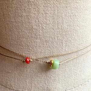 May include: A delicate layered necklace with a small red bead and a larger green bead. The necklace is made of thin brown cord.