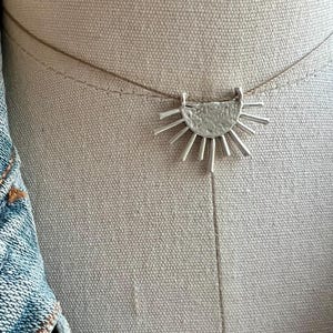May include: A silver-colored necklace with a sunburst pendant. The pendant has a textured, semi-circular design with radiating lines. The necklace is on a thin, neutral-colored cord, and is displayed against a neutral background.