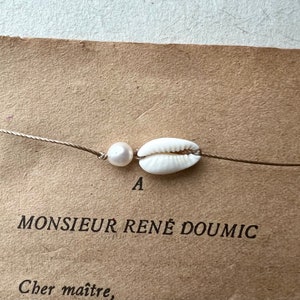 May include: A delicate necklace featuring a white cowrie shell and a pearl, strung on a thin, neutral-colored cord. The necklace is set against a vintage paper background with the text "MONSIEUR RENE DOUMIC".