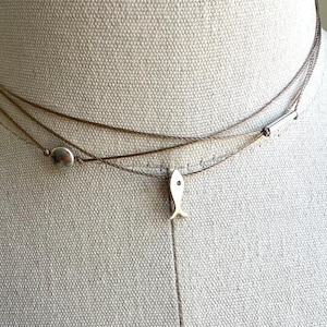 May include: A layered necklace with three strands. The top strand is a silver chain, the middle strand is a thin brown cord with a silver disc, and the bottom strand has a small silver fish pendant with a black dot eye.