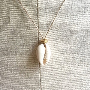 May include: A white cowrie shell pendant necklace with a gold bead on a natural twine cord.