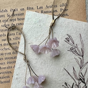May include: Delicate floral earrings featuring pale lavender flower charms suspended from gold-toned wire hoops. The earrings are set against a backdrop of vintage paper with French text and botanical illustrations.