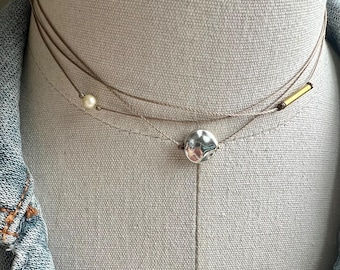 May include: Delicate layered necklace with a silver hammered disc pendant, a small pearl, and a gold-toned bar. The necklace is displayed on a neutral-toned mannequin, showcasing its minimalist design and elegant style.